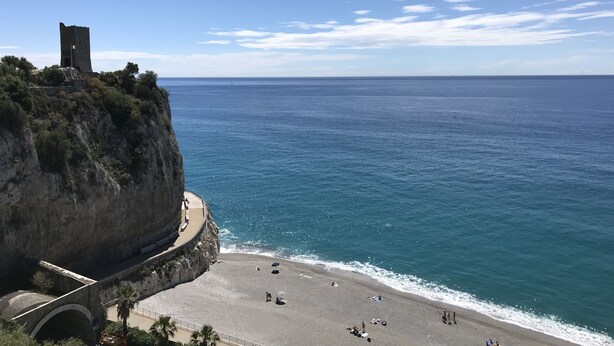 Eine Küstenansicht mit einem historischen Turm auf einer Klippe, einem Strand mit Menschen und einem klaren Himmel.