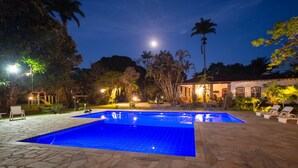 Uma área de piscina com vista desobstruída para a lua no céu noturno, cercada por palmeiras e um edifício com telhado de azulejos.