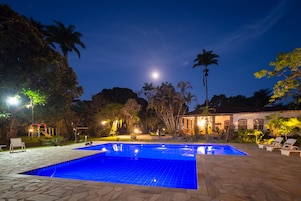Uma área de piscina com vista desobstruída para a lua no céu noturno, cercada por palmeiras e um edifício com telhado de azulejos.