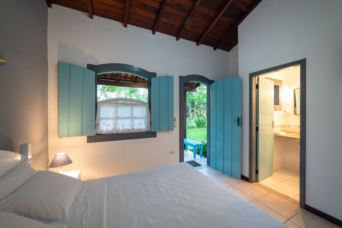 A bedroom with a bed, a window with curtains, a nightstand with a lamp, and a view of the garden through the window.