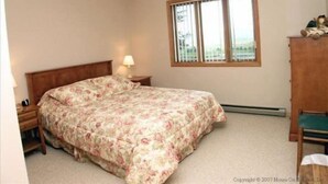 A bedroom with a single bed, a nightstand, a lamp, a window with blinds, and a view of the outdoors.