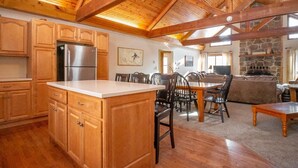 A spacious kitchen with wooden cabinets and a large island, a dining area with chairs, and a stone fireplace in the living room. - Mountain Treat (Davis)