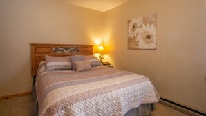 A bedroom with a wooden headboard, a bed with a striped blanket, a bedside lamp, and a floral painting on the wall. - Mountain Treat (Davis)