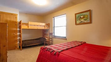 A dormitory room with bunk beds, a single bed, a wooden wardrobe, a window with blinds, and a painting on the wall.