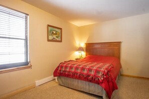 Un dormitorio con cama, ventana con persianas, lámpara y un cuadro en la pared.