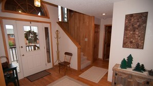 Una escalera de madera que conduce a un segundo piso, una puerta con panel de vidrio, una silla y una pequeña mesa con una cesta.
