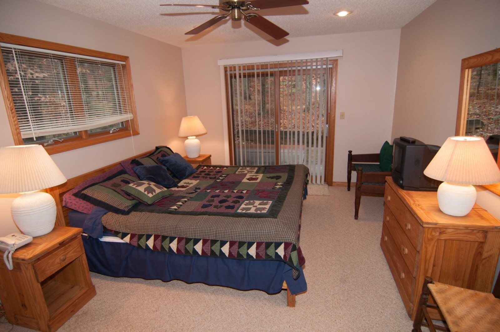 A bedroom with a large bed, two bedside tables with lamps, a wooden dresser, a chair, a ceiling fan, and a window with blinds.