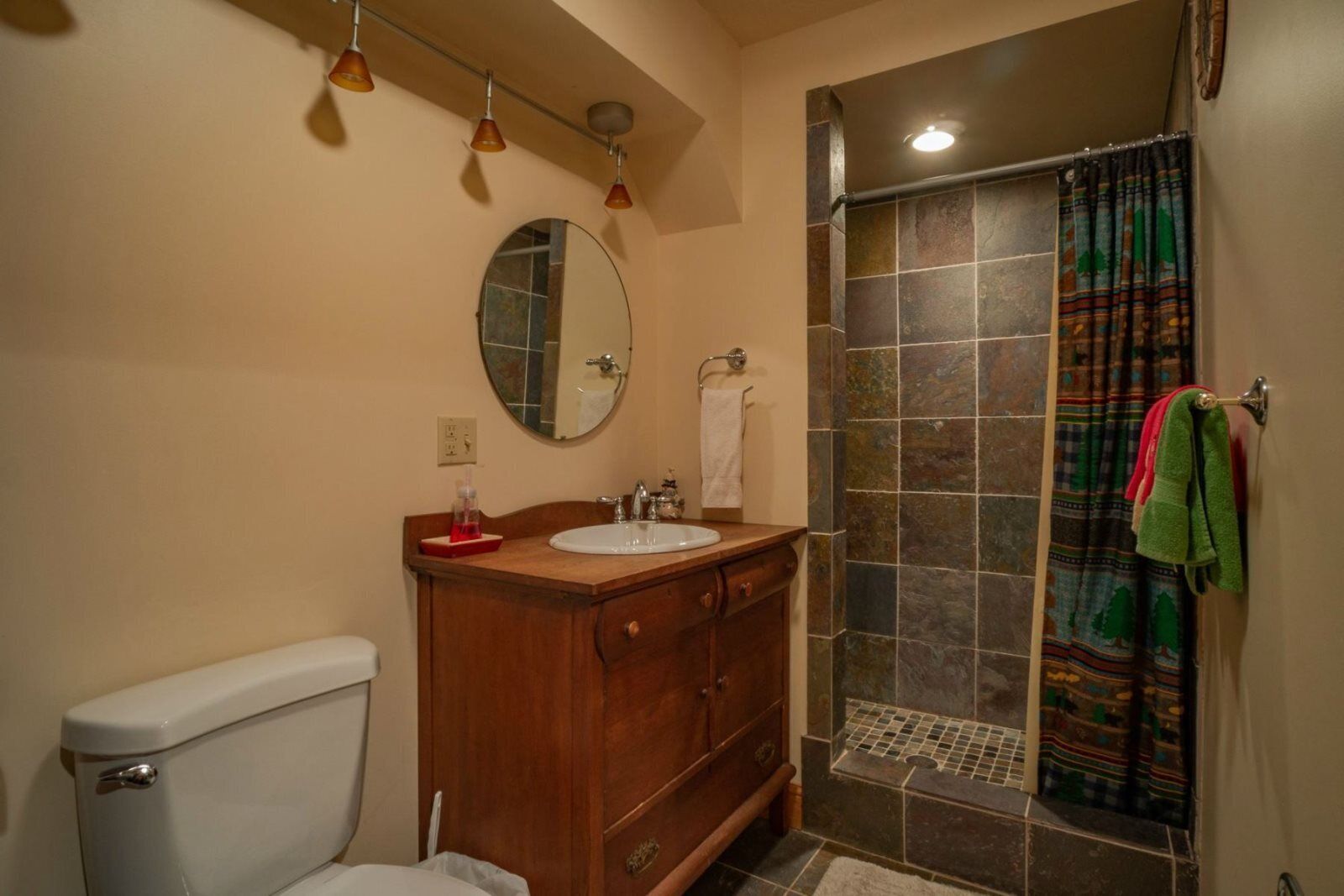 A bathroom with a wooden vanity, a white toilet, a shower with a tiled wall, and a mirror above the sink.