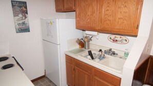 A compact kitchen with wooden cabinets, a white refrigerator, a sink with various utensils, and a poster on the wall. - Herzwoods D304 (Davis)