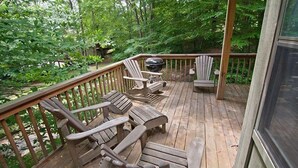 Une terrasse en bois, agrémentée de chaises Adirondack et d’un barbecue, le tout entouré d’une végétation luxuriante.