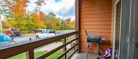 A wooden deck with a barbecue grill, a concrete floor, and a view of a parking lot with vehicles and autumn trees.