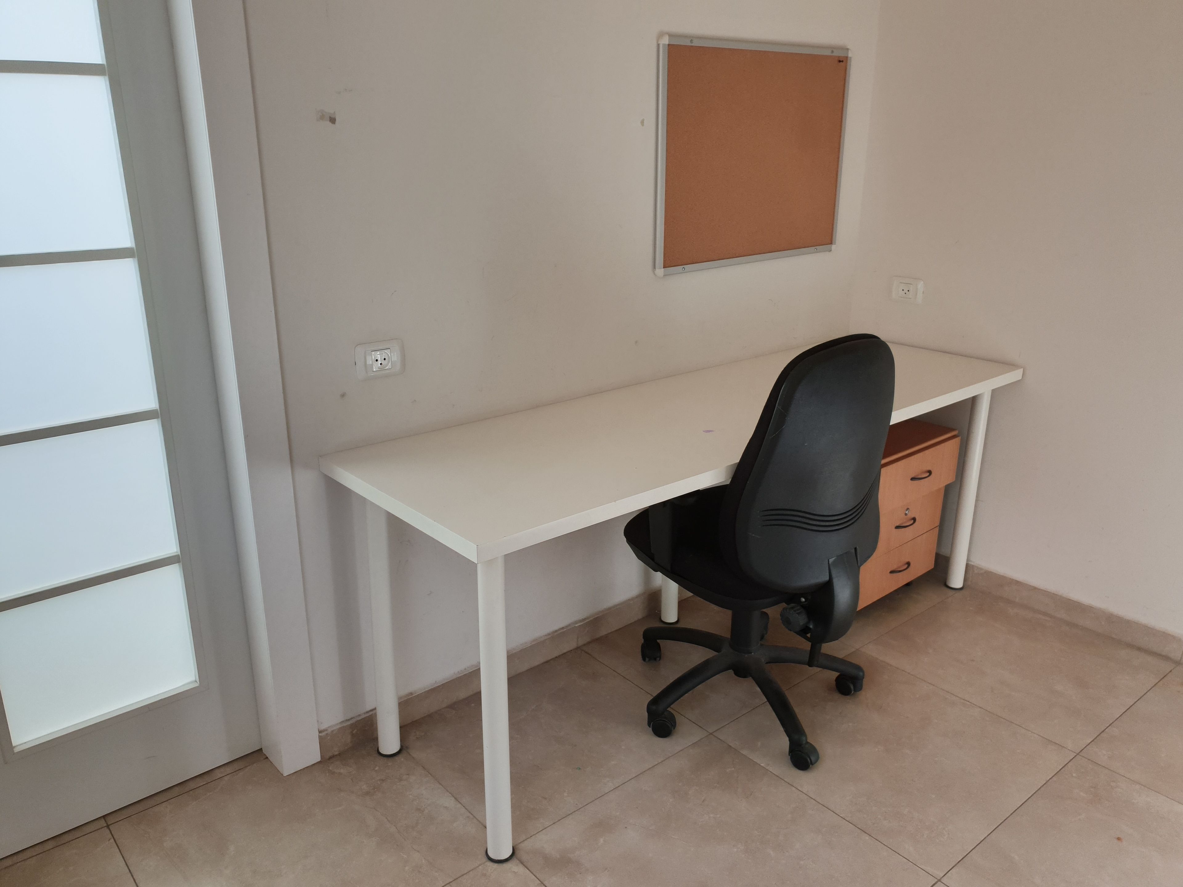 A white desk with a black office chair, a corkboard on the wall, and a wooden drawer unit.