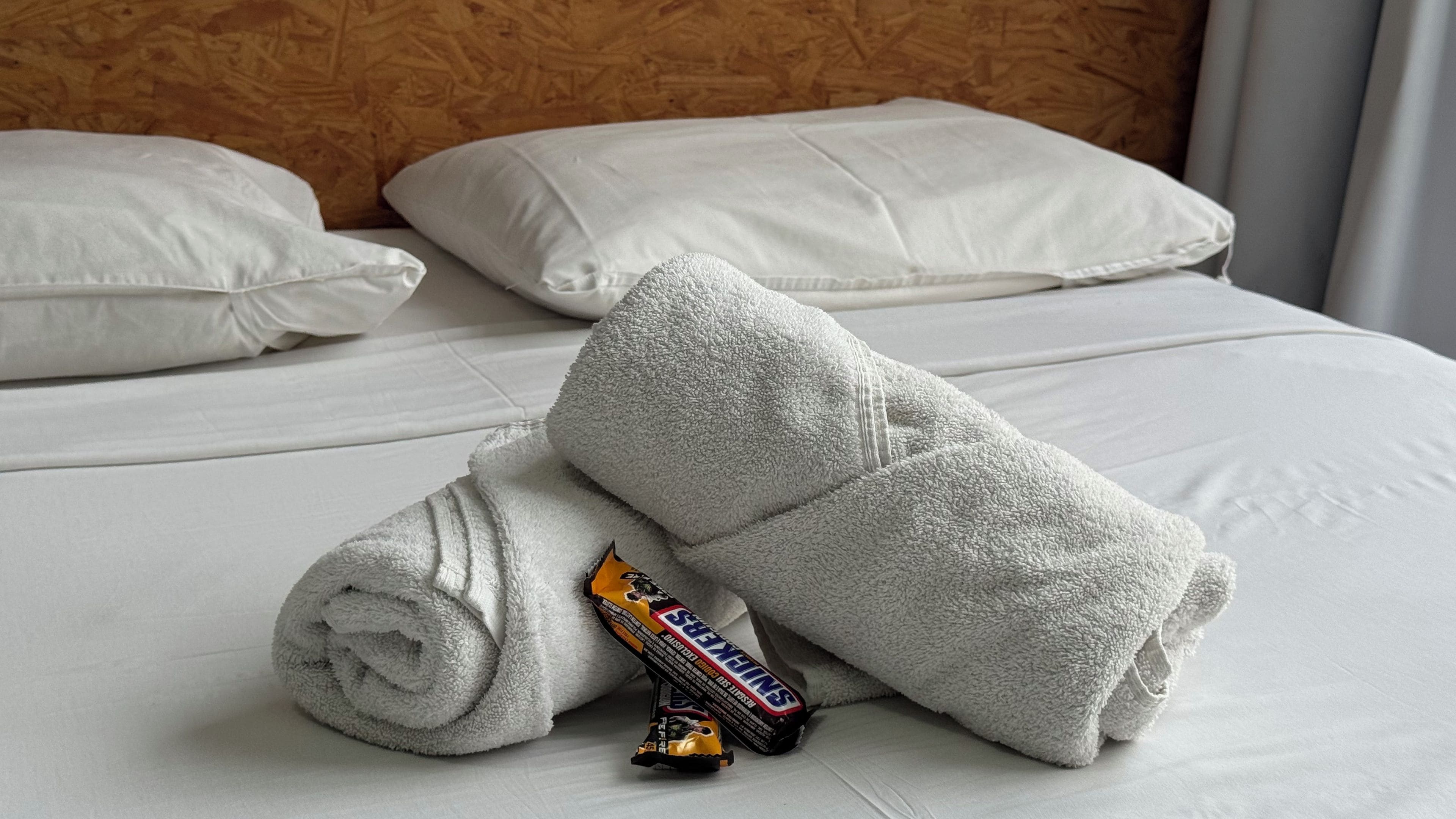 A neatly made bed with white linens and pillows, a folded towel, and a Snickers chocolate bar.