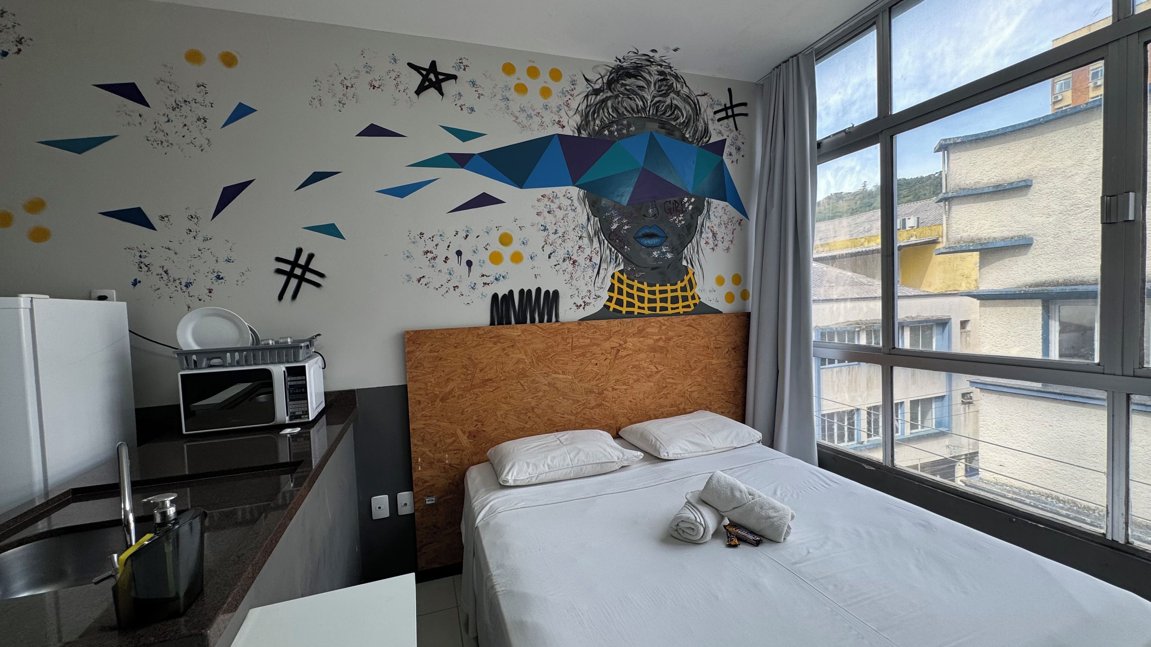 Une chambre d’hôtel avec un lit, un bureau doté d’un micro-ondes, une fenêtre donnant sur des immeubles et un mur orné d’une fresque géométrique.