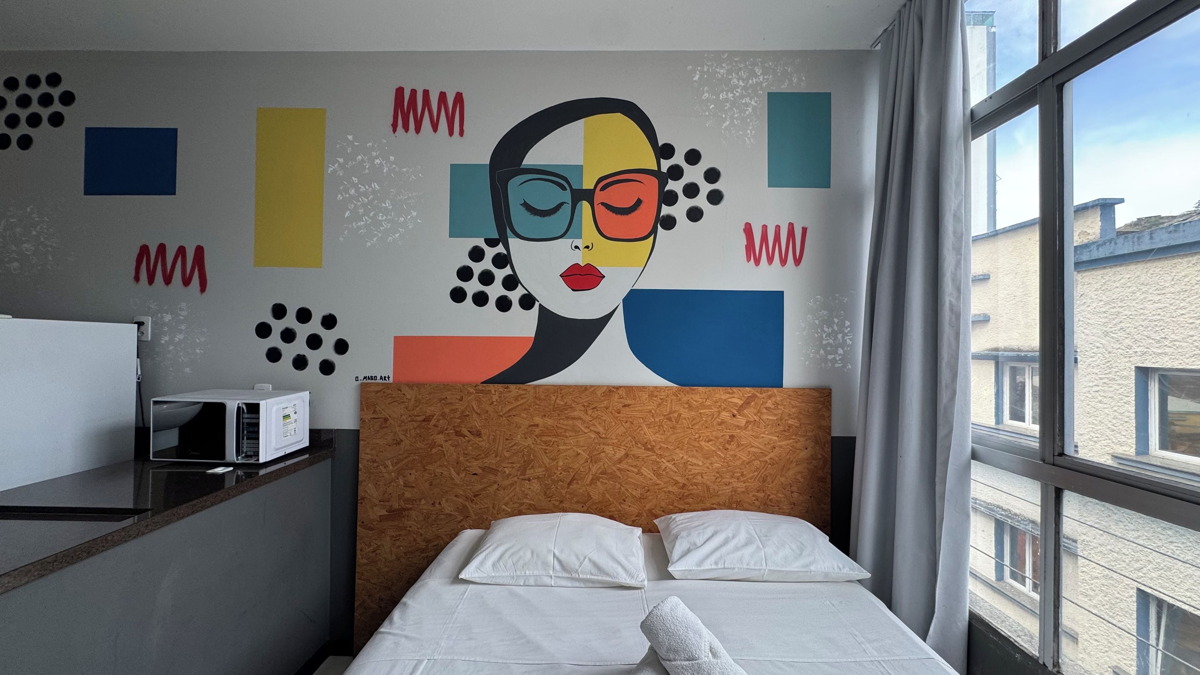 Habitación de hotel con una cama, cabecero de madera, pared con arte abstracto, ventana y escritorio con microondas.