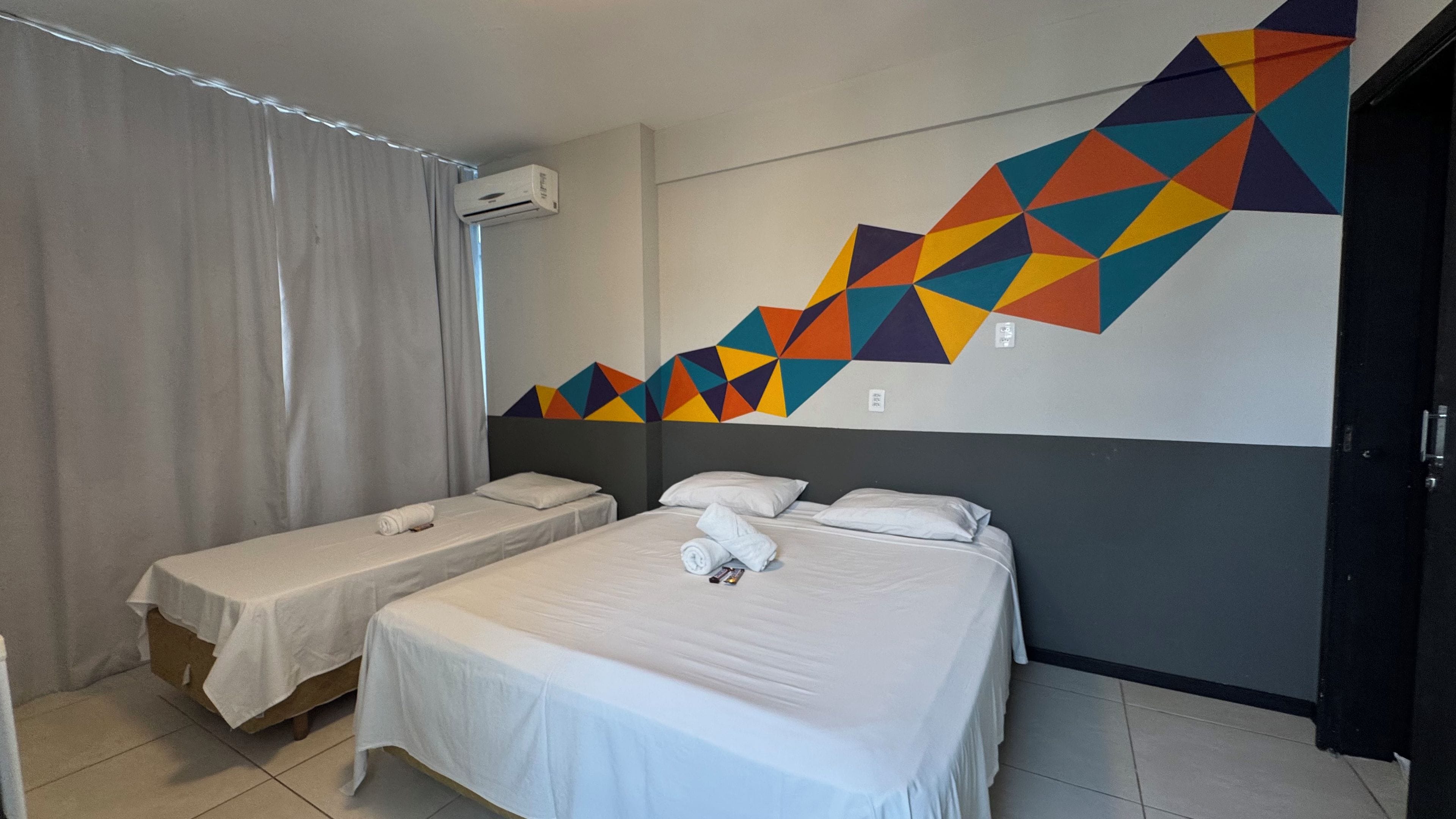 A hotel room with two beds, a window with curtains, an air conditioning unit, and a colorful geometric wall design.