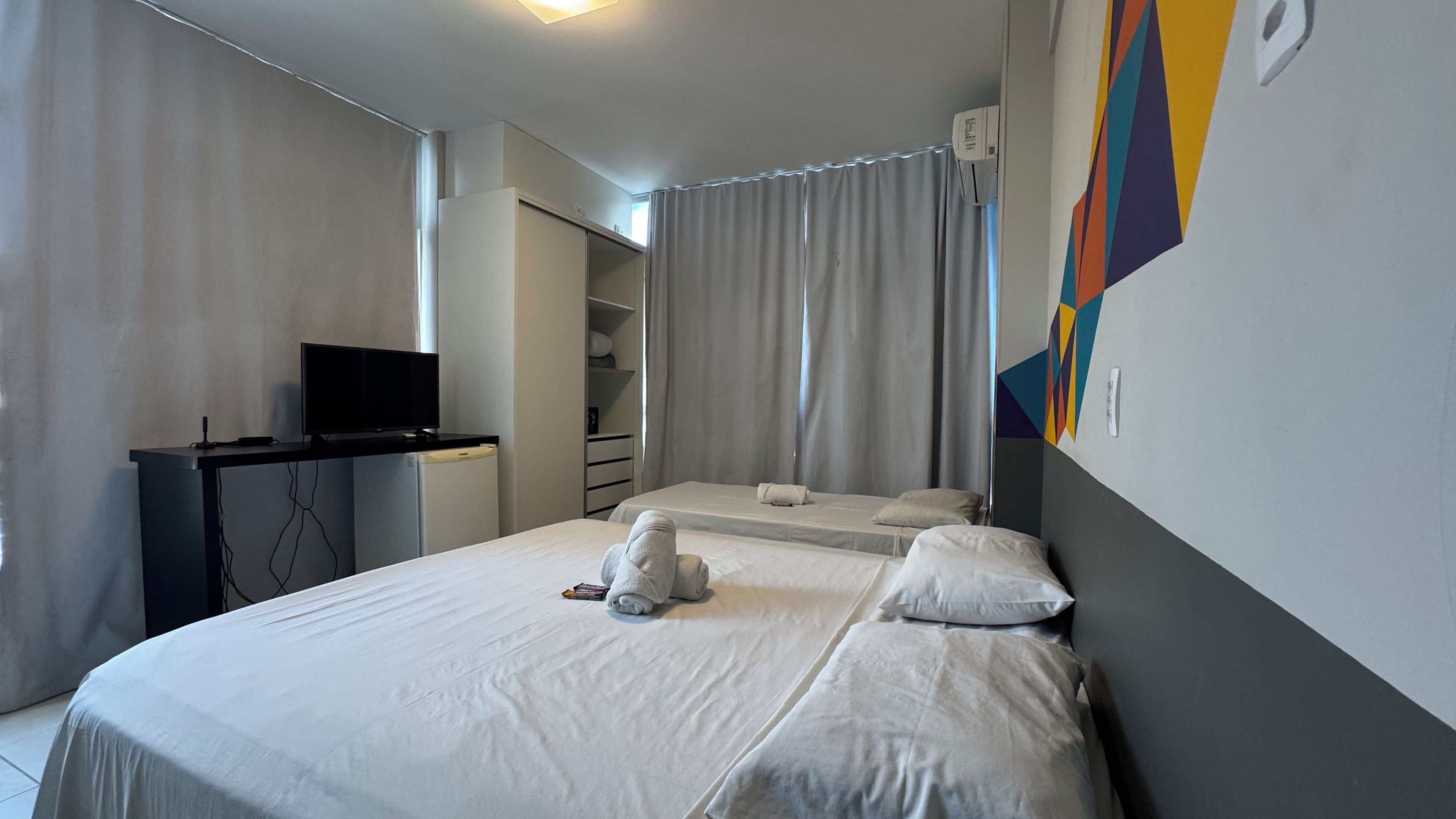 A hotel room with a bed, a desk with a television, a wardrobe, and a wall-mounted air conditioning unit.