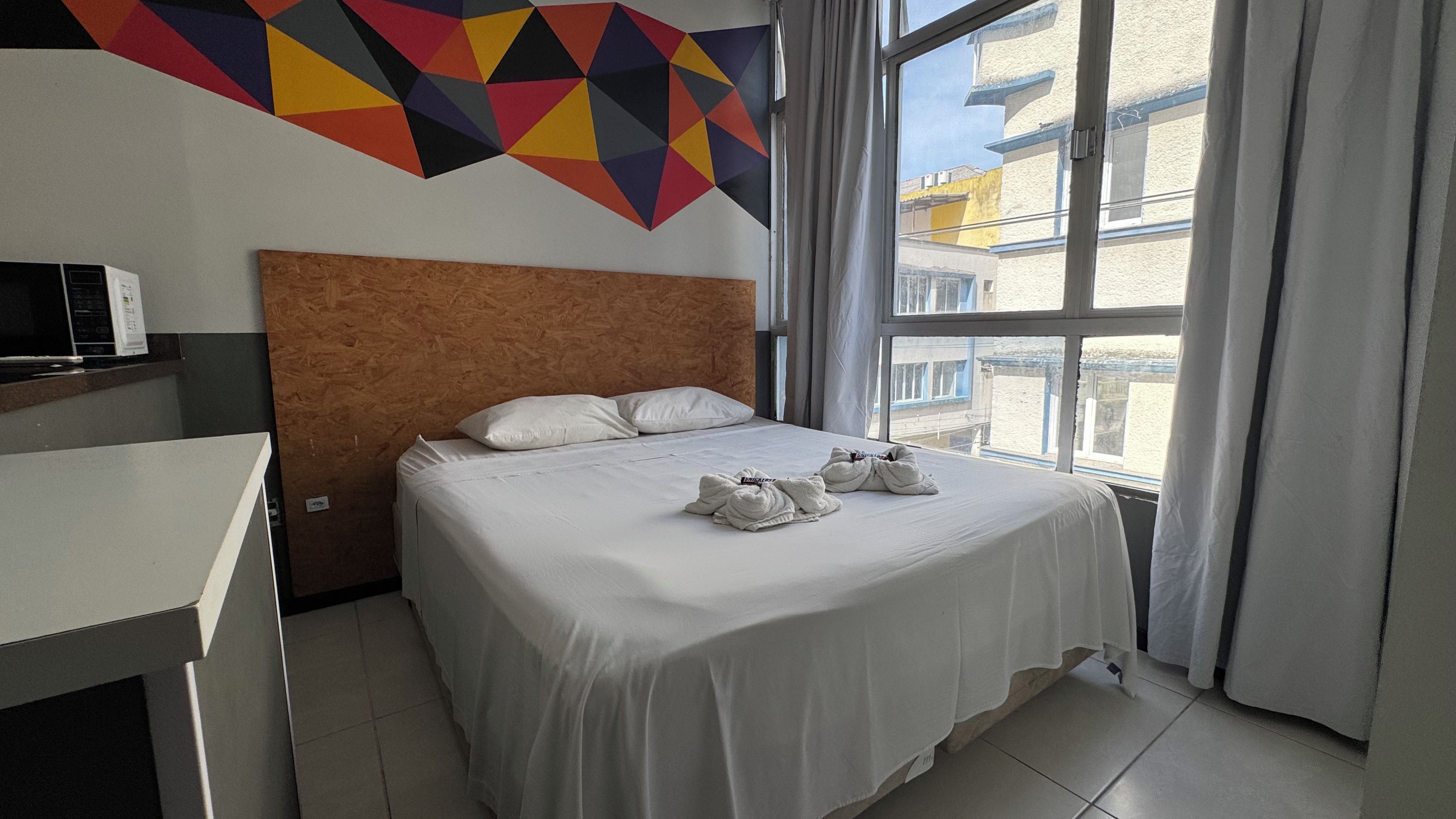 A hotel room with a bed, pillows, a headboard, a window with curtains, and a geometric wall decoration.