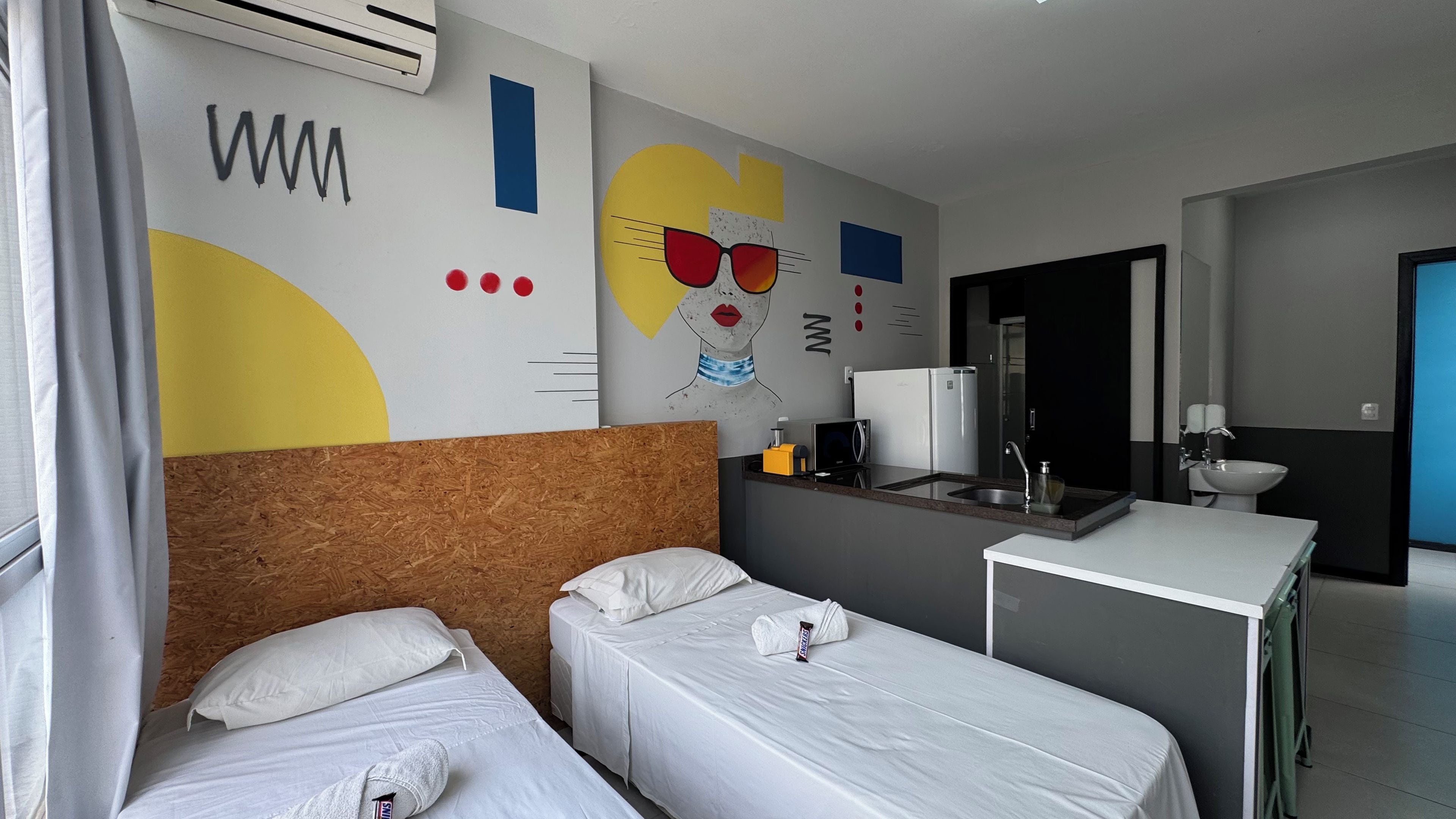 Une chambre d’hôtel avec deux lits simples, une petite kitchenette, un miroir et une salle de bain équipée d’un lavabo et de toilettes.