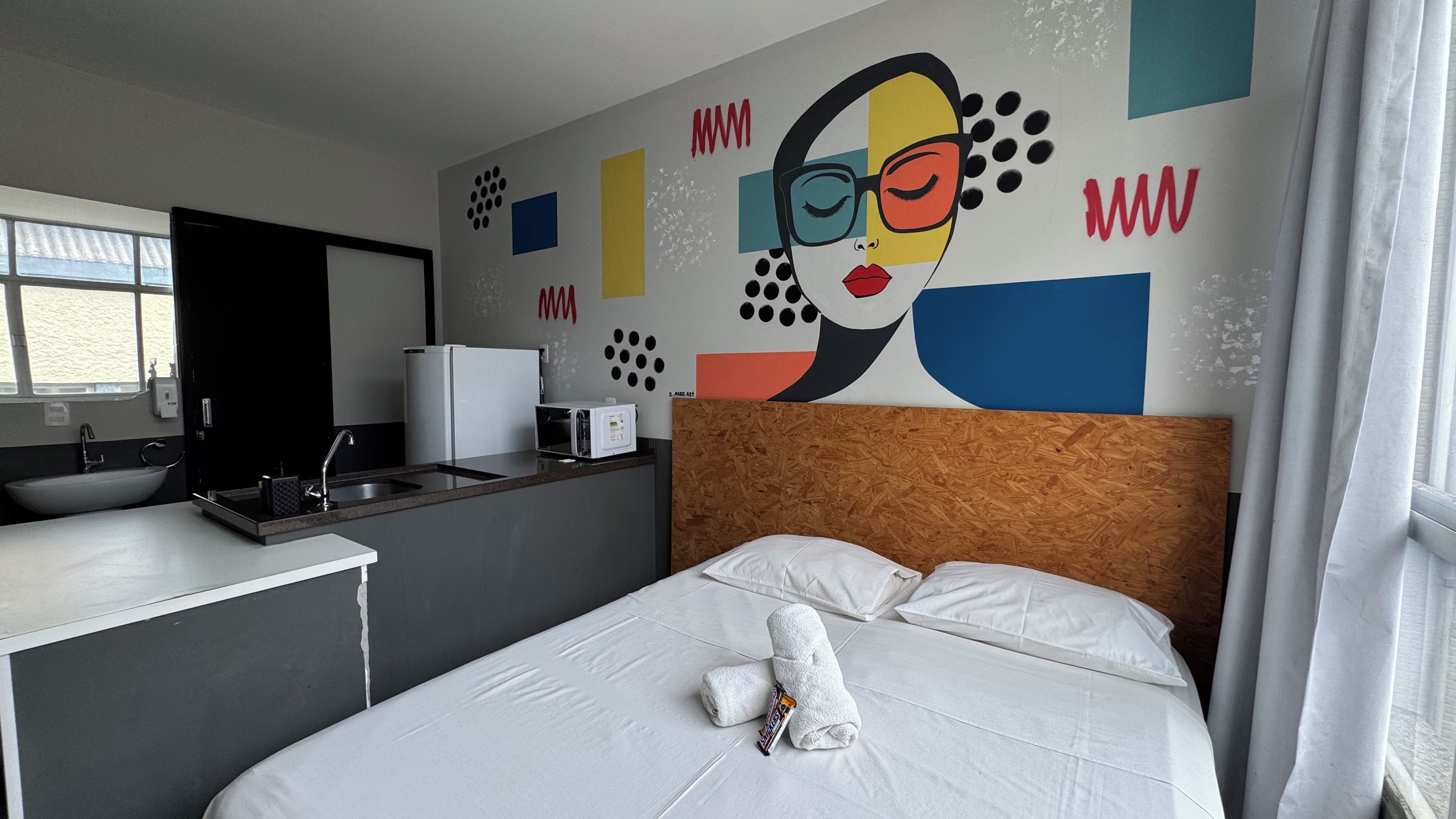 A hotel room with a bed, a small kitchenette, a sink, and a window.