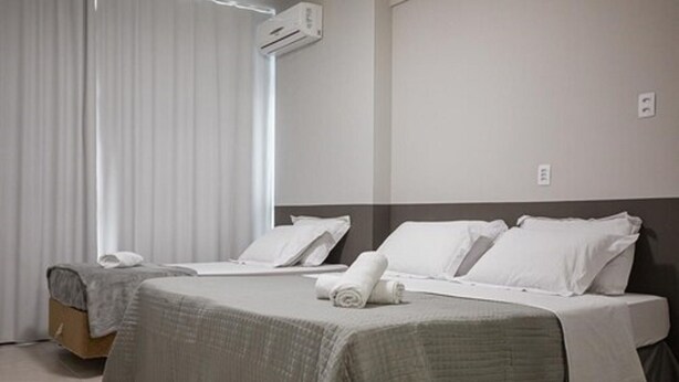Hotelkamer met twee bedden, elk voorzien van witte lakens en een grijze plaid. Er is een raam met witte gordijnen, een airconditioningunit aan de wand en een houten vloer.