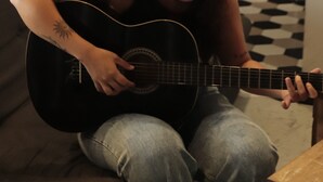 Een persoon speelt op een gitaar op een bank in een kamer met een geruite vloer en een koelkast op de achtergrond.