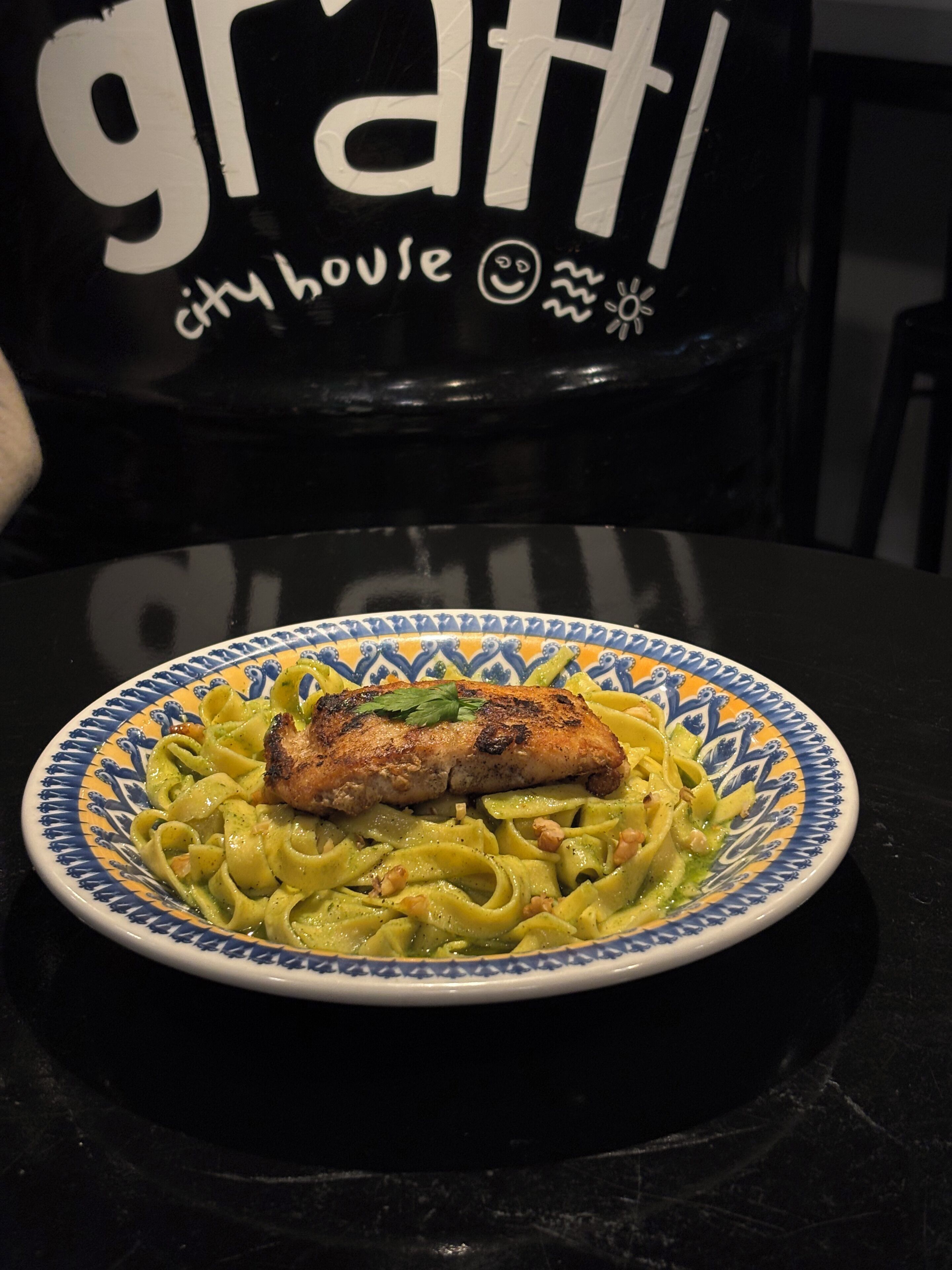 A plate of pasta with a piece of grilled chicken on top, placed on a table with a black chair in the background.