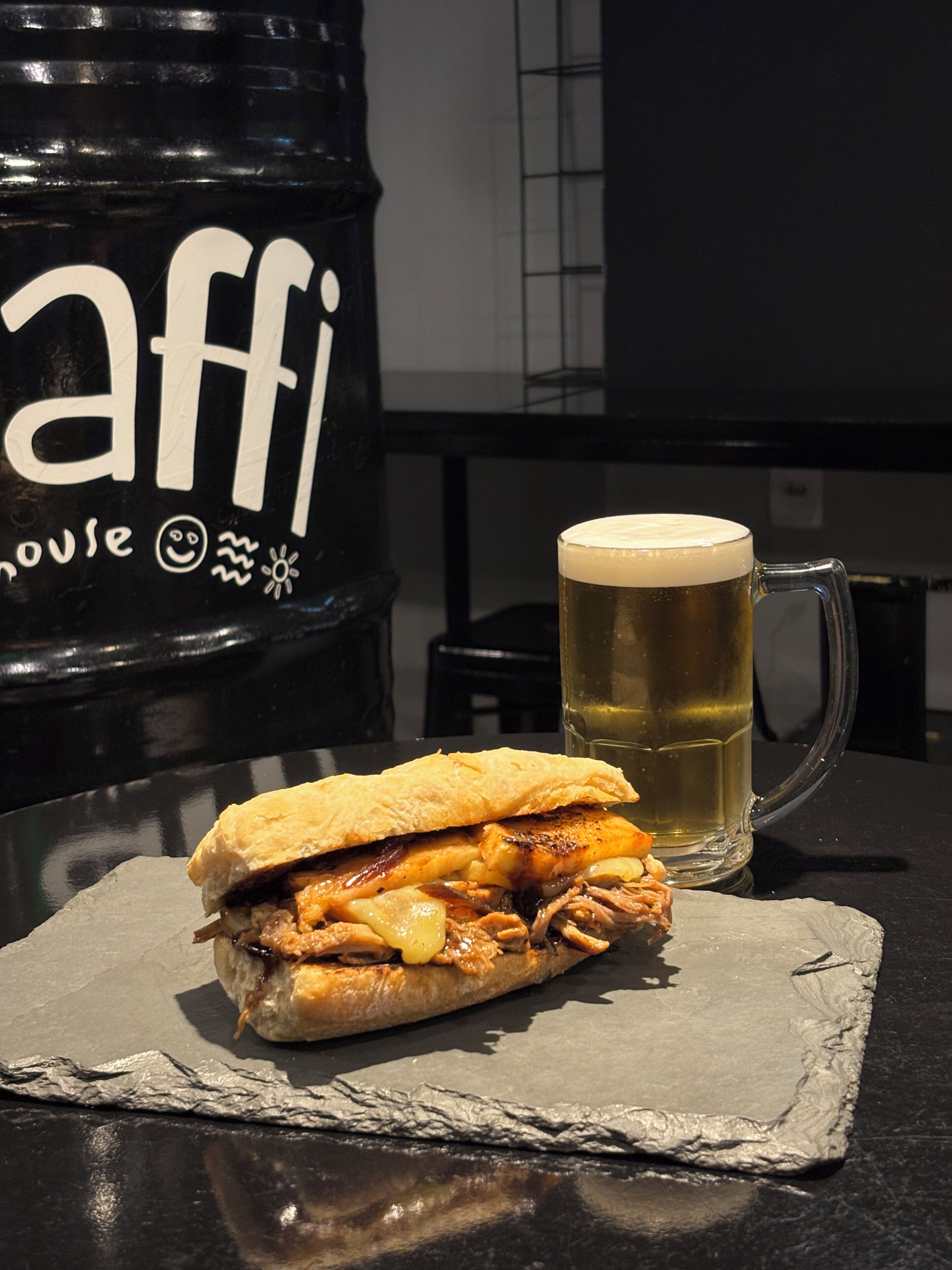A sandwich on a slate coaster, a mug of beer, and a black container with white text.