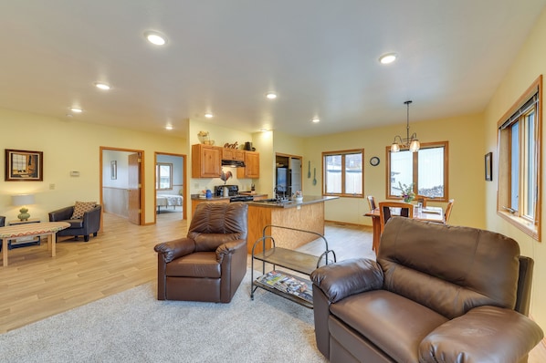 A spacious living room with leather sofas, a dining area, a kitchen with wooden cabinets, and a large window providing natural light.