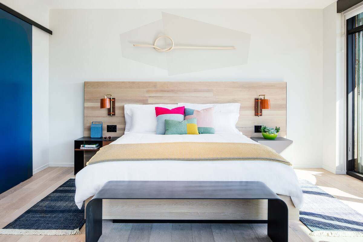 A modern bedroom with a wooden headboard, a bed with multiple pillows, bedside tables with lamps, and a large window providing natural light.