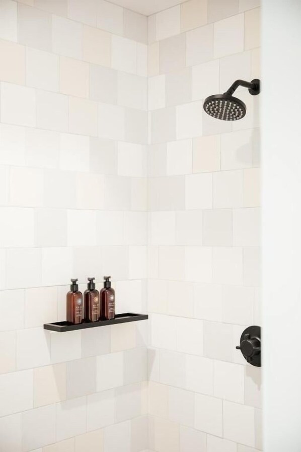 Zona de ducha con azulejos blancos, un cabezal de ducha negro y un estante con tres botellas.