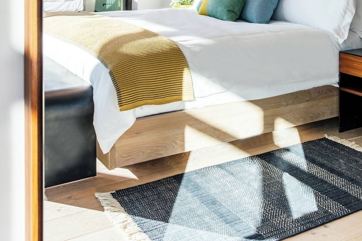 A modern bedroom with a large bed, a wooden headboard, a colorful pillow arrangement, and a view through a glass door.