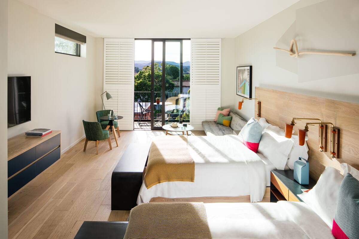 A modern hotel room with a large bed, a desk, a chair, a balcony with a view of the mountains, and a TV.