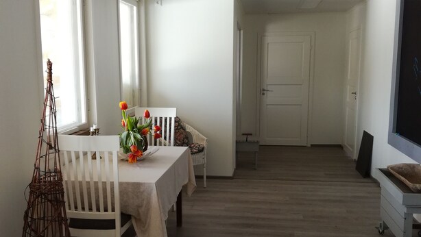 Une pièce spacieuse avec une table à manger dressée pour quatre personnes, une chaise blanche et une petite table sur laquelle est posé un vase de fleurs.