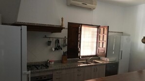 Una cocina con estufa a gas, un lavamanos y alacenas. Hay una ventana con persianas y una repisa para colgar ollas.