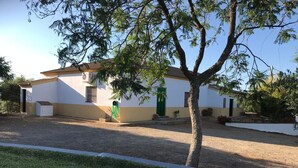 Una casa de una sola planta con fachada blanca y puerta verde, rodeada por un camino de grava y un césped.