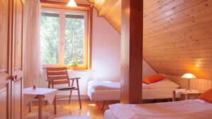 A cozy bedroom with a wooden ceiling, a window with a view of trees, a small table with a chair, and a bedside lamp.