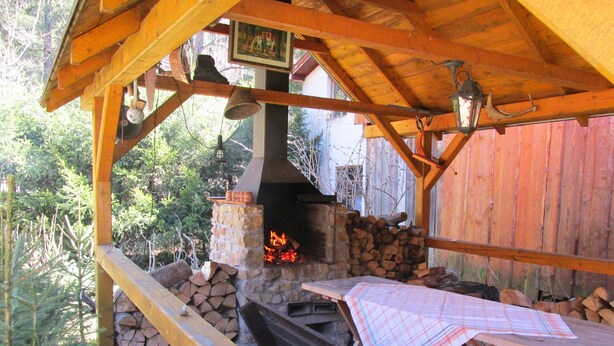 A rustic outdoor kitchen with a stone oven, wooden beams, and a wooden table with a plaid tablecloth.
