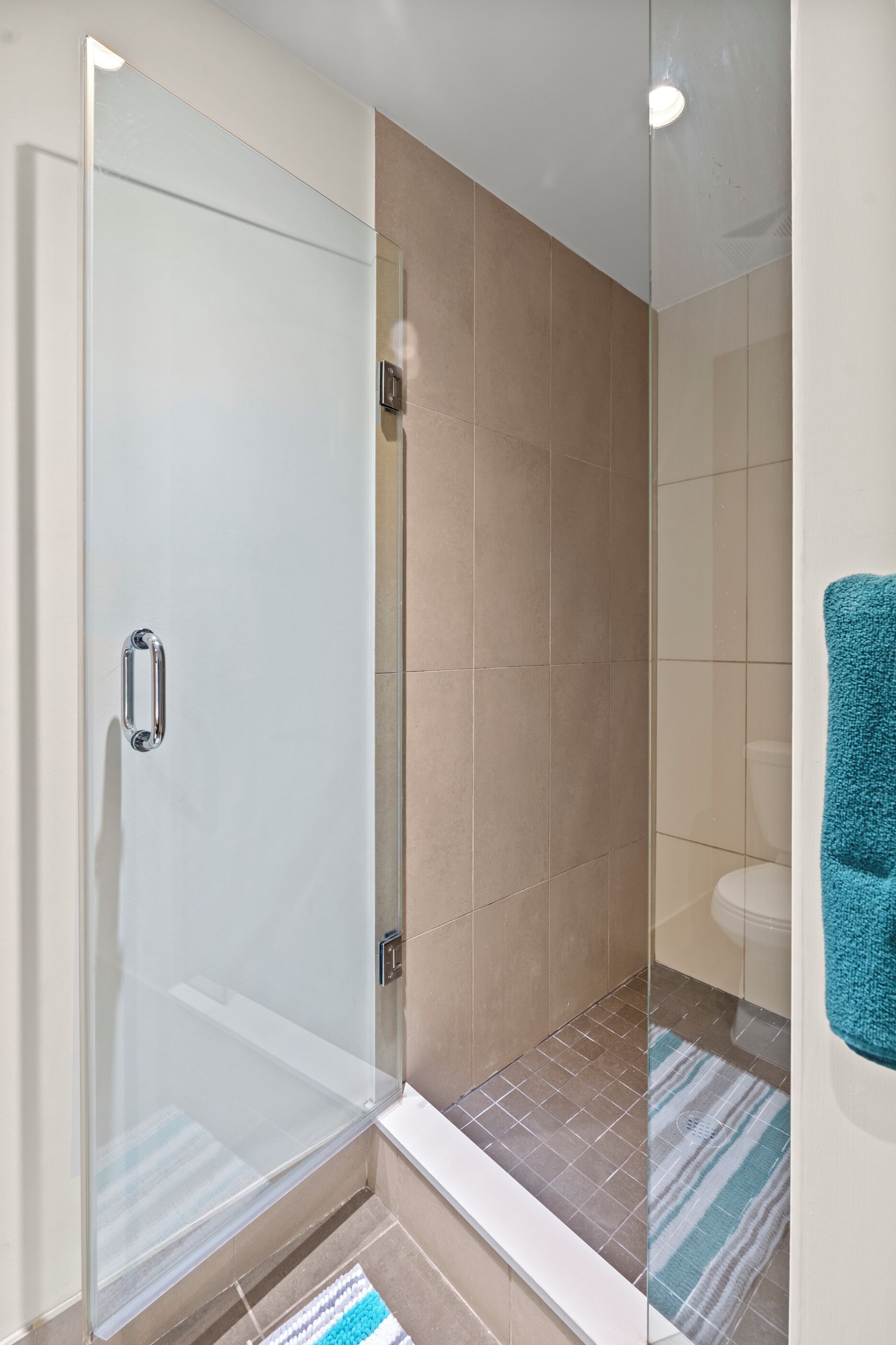 A modern bathroom with a glass shower enclosure, beige tiles, and a striped bath mat.