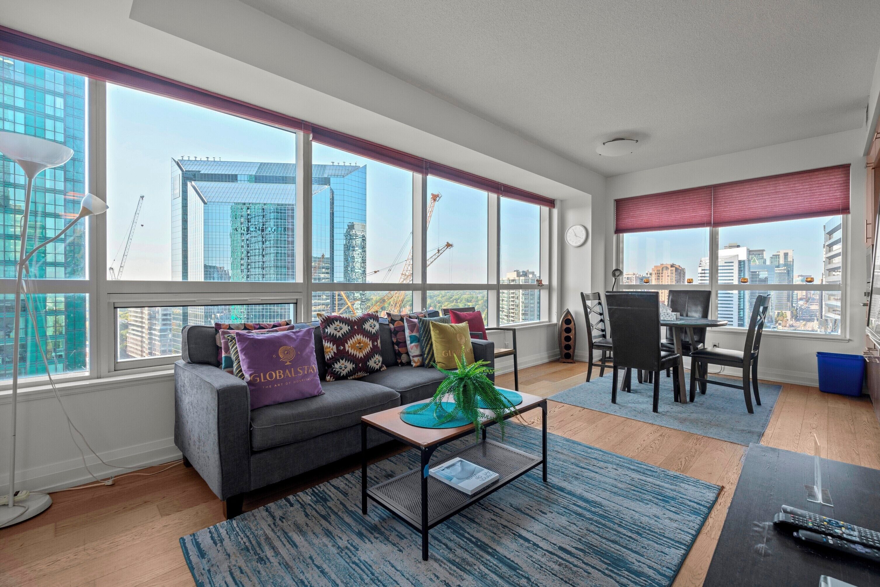 Sala de estar moderna com sofá cinzento, almofadas coloridas, mesa de centro e área de jantar com mesa e cadeiras. Grandes janelas oferecem vista para o horizonte da cidade e guindastes de construção.