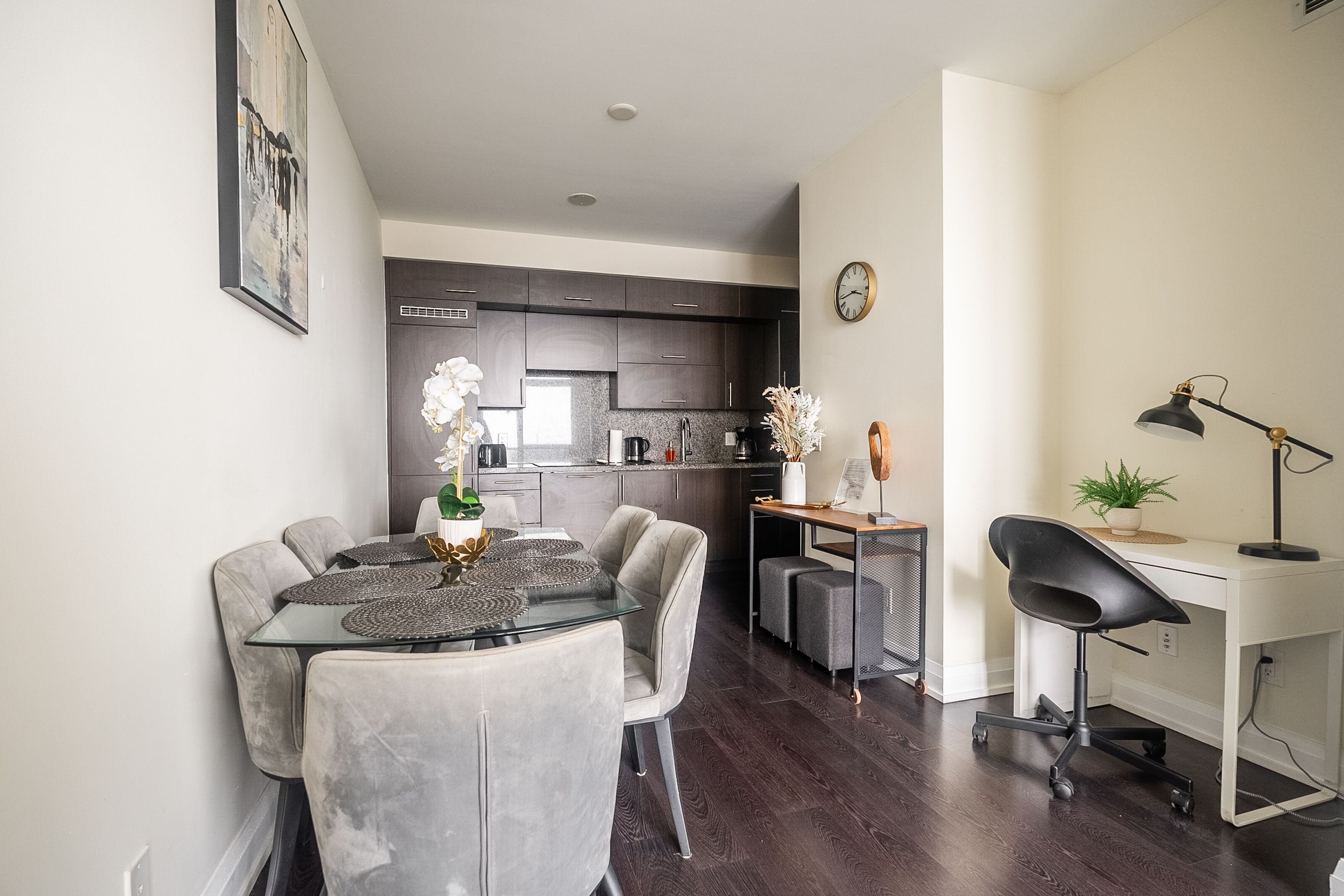 A modern kitchen with dark cabinets, a dining area with a glass table and grey chairs, and a desk with a chair and a lamp.