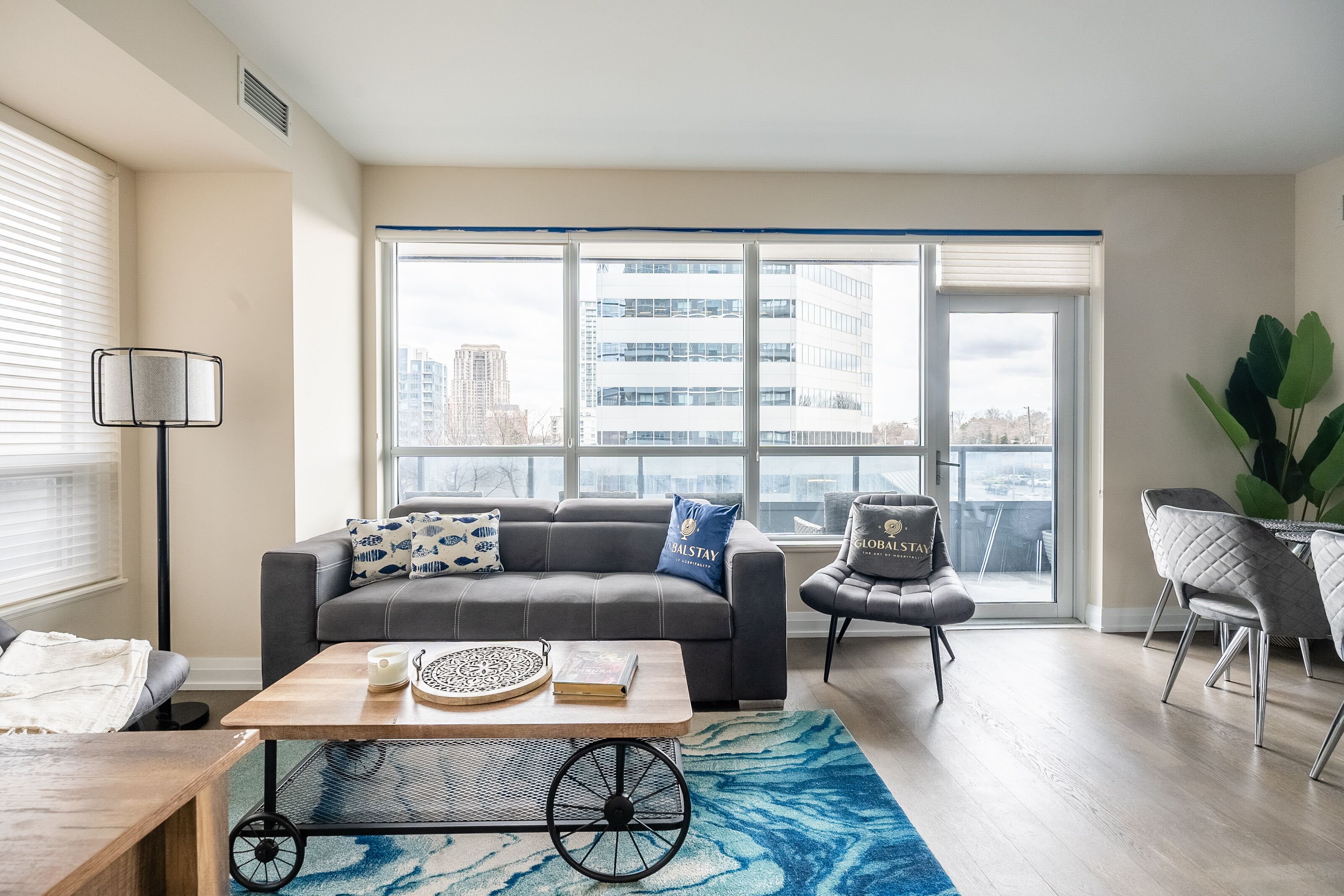 A modern living room with a grey sofa, a coffee table, and a view of a cityscape through large windows.