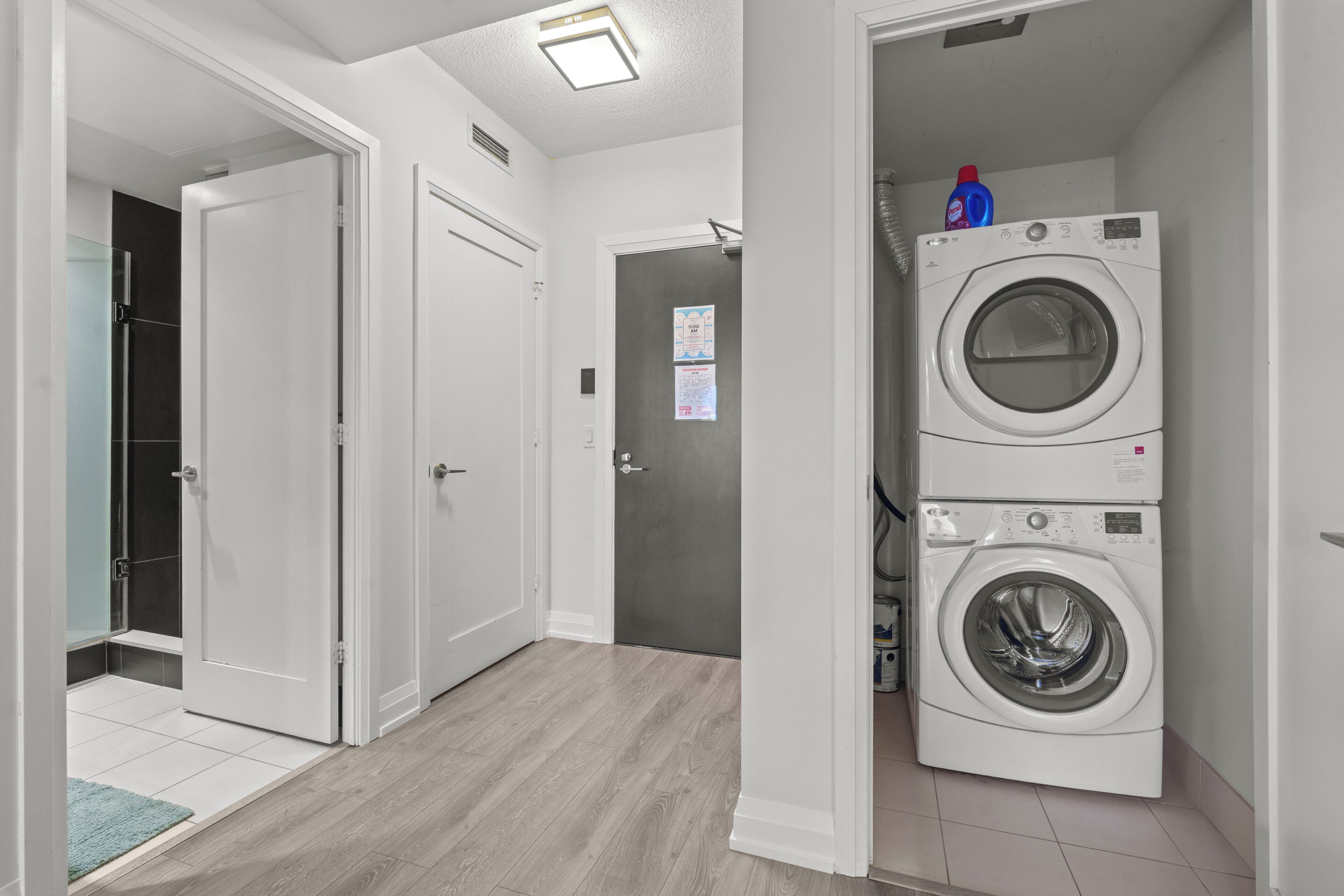 A laundry area with a front-loading washer and dryer, a door leading to a bathroom, and a hallway with a light fixture.