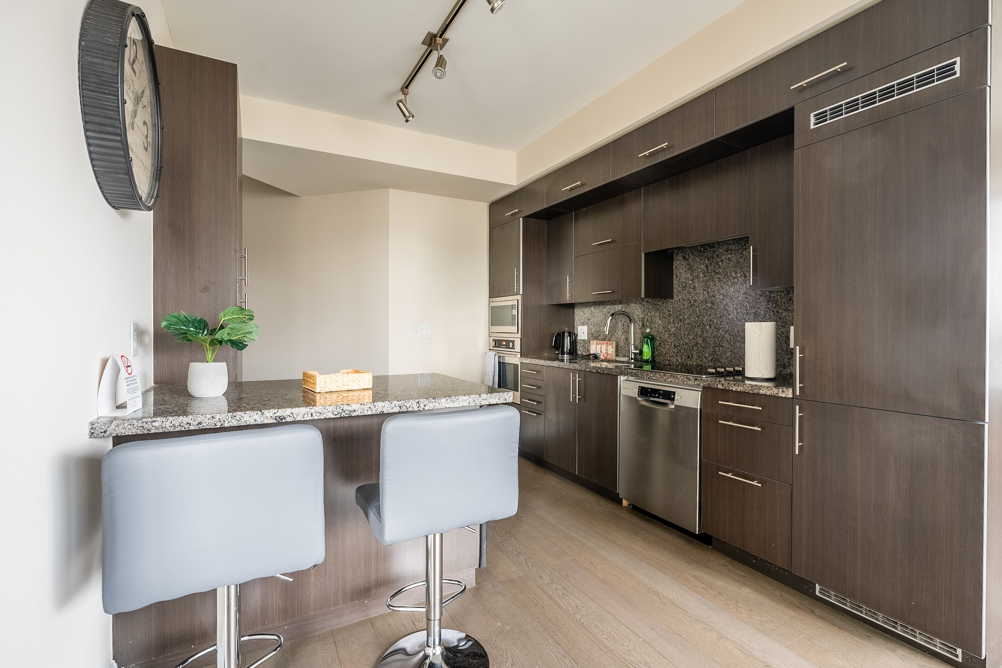 A modern kitchen with dark wood cabinets, a granite countertop, and stainless steel appliances. There are two bar stools and a potted plant on the counter.