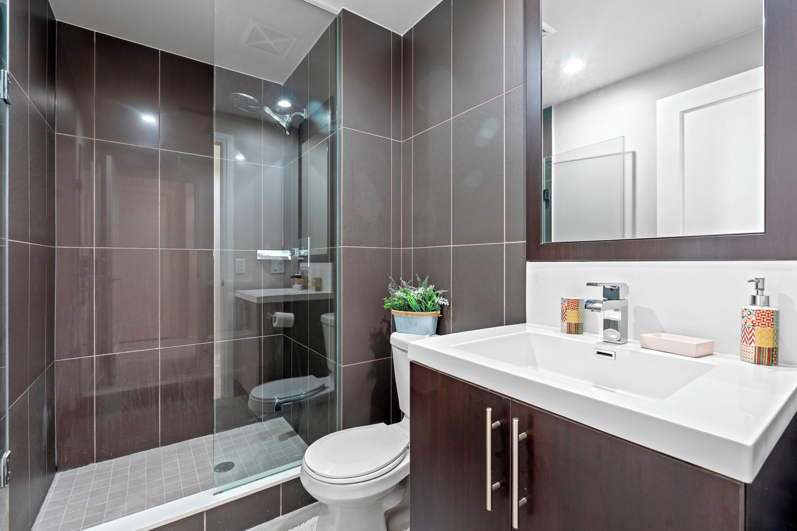 A modern bathroom with dark tiles, a glass shower enclosure, a white toilet, and a vanity with a mirror.