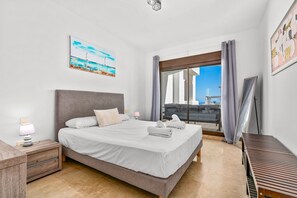 Een moderne hotelkamer met een bed, nachtkastje, lamp en uitzicht op zee door een groot raam.