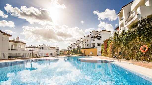 En swimmingpool omgivet af hvide bygninger med altaner, en klar blå himmel og en redningskrans til højre.
