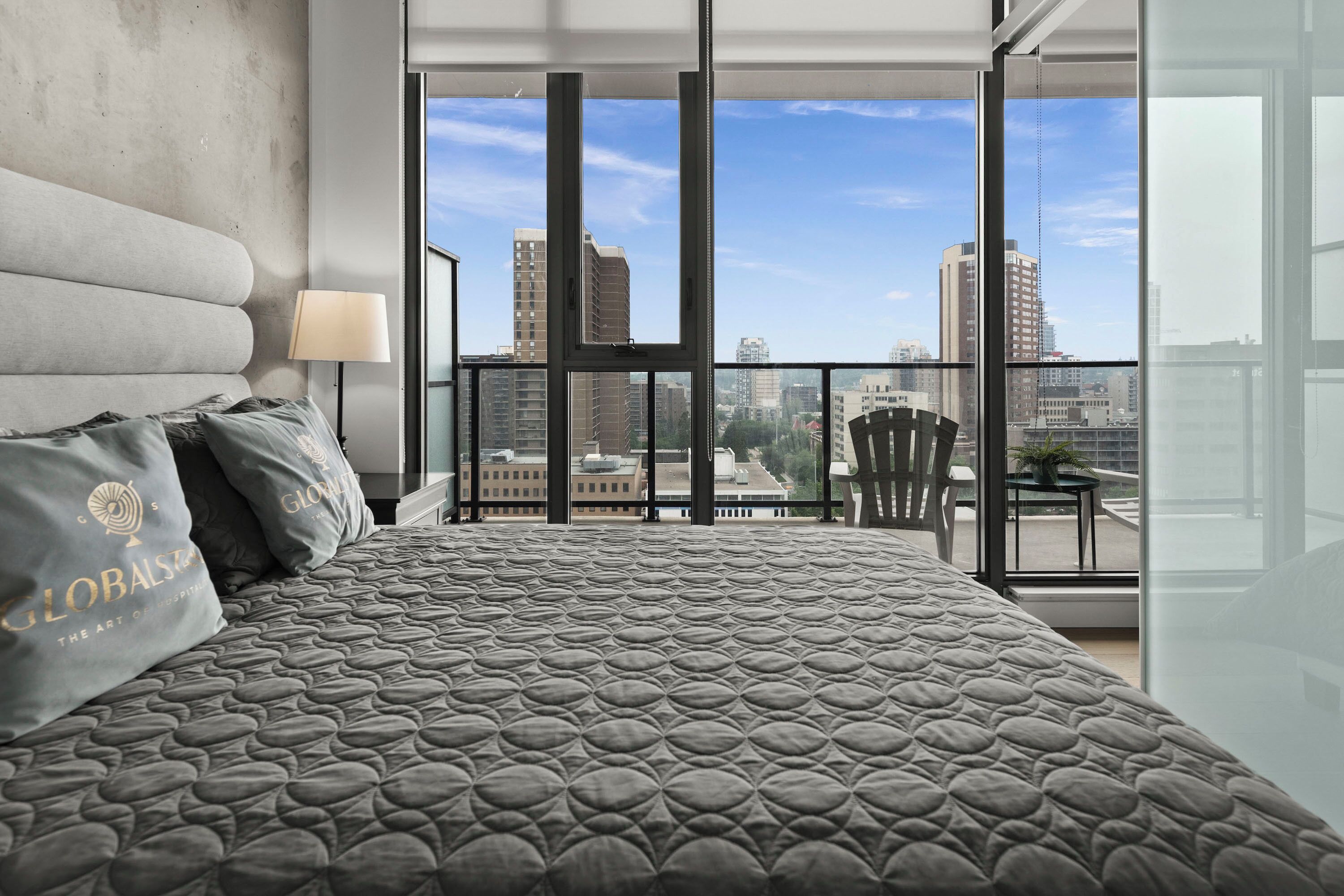 Une chambre moderne dotée d’un grand lit, offrant une vue sur la ville grâce à de grandes fenêtres du sol au plafond, et d’un balcon avec une chaise et une table.