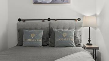 Una cama bien hecha con ropa de cama gris y dos almohadas con la marca 'GLOBALSTAY'. Se ve una mesita de noche con una lámpara a la derecha.
