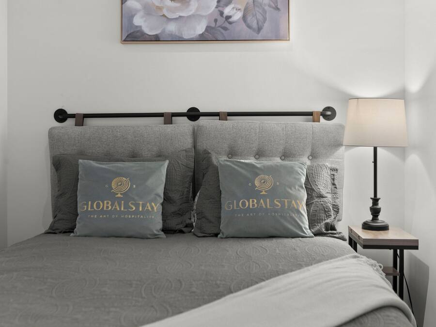 Una cama bien hecha con ropa de cama gris y dos almohadas con la marca 'GLOBALSTAY'. Se ve una mesita de noche con una lámpara a la derecha.